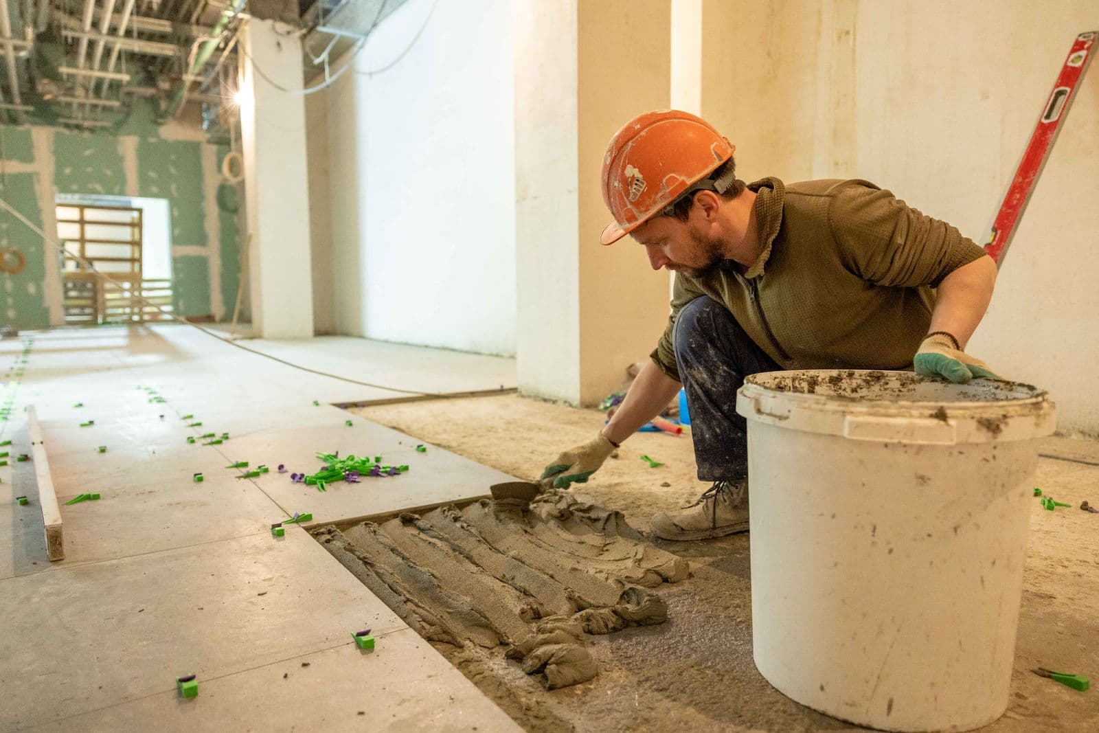 Tiler kneeling on a protected floor, combing adhesive and bedding a large floor tile